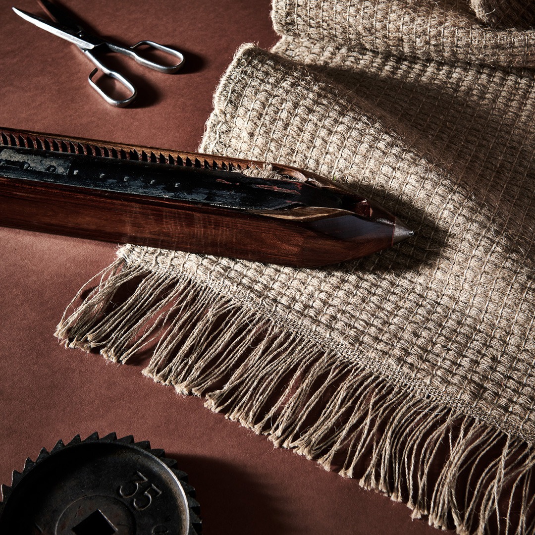 A close-up of a linen woven rug with fringes on a brown surface, accompanied by a pair of scissors and a wooden weaving shuttle.